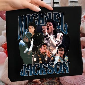 Puede incluir: Bolso tote negro con un collage de imágenes de Michael Jackson. El nombre "MICHAEL JACKSON" está arqueado en la parte superior en azul, con "JACKSON" debajo. El diseño tiene una estética vintage y retro.