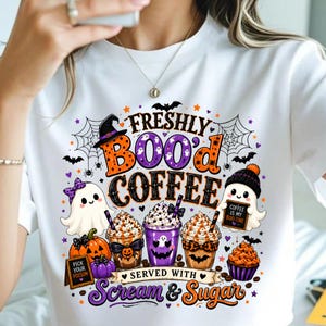Puede incluir: Una camiseta blanca con un gráfico colorido de Halloween con el texto "FRESHLY BOO'D COFFEE SERVED WITH SCREAM & SUGAR". El diseño incluye fantasmas, calabazas, tazas de café, telarañas, murciélagos y un pequeño letrero que dice "POISON".