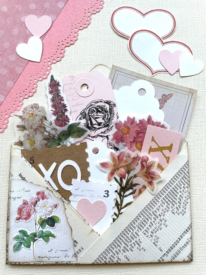 以下が含まれることがあります： ピンクと白の花柄のヴィンテージ封筒。紙のハート、花、白い「XO」の文字で飾られています。封筒は一部開いており、手書きの文字が書かれた茶色の内側が見えています。