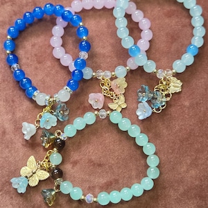 Puede incluir: Cuatro pulseras de cuentas en tonos azul, rosa y verde. Cada pulsera presenta cuentas redondas y dijes decorativos, incluyendo flores y mariposas. Las pulseras se exhiben sobre una superficie marrón.