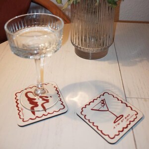 May include: A clear coupe glass with liquid sits on a white coaster with a red border and a cocktail illustration. A second coaster with a similar design is next to it. A vase of dried flowers is in the background.