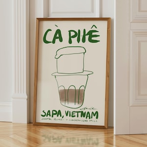 May include: A framed art print featuring a green illustration of Vietnamese coffee with the text "C&agrave; Ph&ecirc;" at the top. Below the coffee illustration is the text "Sapa, Vietnam". The print is in a wooden frame.
