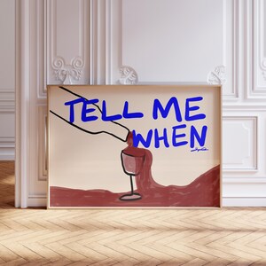 Könnte beinhalten: Ein gerahmter Kunstdruck mit dem blauen Text "TELL ME WHEN", über einer Illustration eines Weinglases und gießenden Weins. Das Kunstwerk hat einen beigen Hintergrund und einen Holzrahmen. Die Kunst wird in einem Raum mit weißen Wänden und Holzboden ausgestellt.