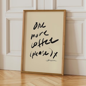 Puede incluir: Un cuadro enmarcado con el texto "One more coffee (please) x" en escritura negra sobre un fondo crema. El marco de madera complementa los tonos neutros de la obra de arte. El cuadro est&aacute; apoyado contra una puerta blanca.