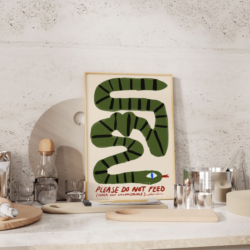 May include: A framed art print with a green snake design and the text "PLEASE DO NOT FEED (UNDER ANY CIRCUMSTANCE)". The snake has black stripes and a red tongue. The artwork is displayed on a white countertop, with other kitchen items in the foreground.