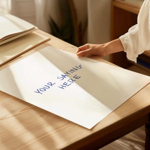 以下が含まれることがあります： 青いインクで「YOUR SAYING HERE」と書かれた白い紙。紙は木製のテーブルの上にあり、手が紙の端を持っています。他の紙が背景にあります。