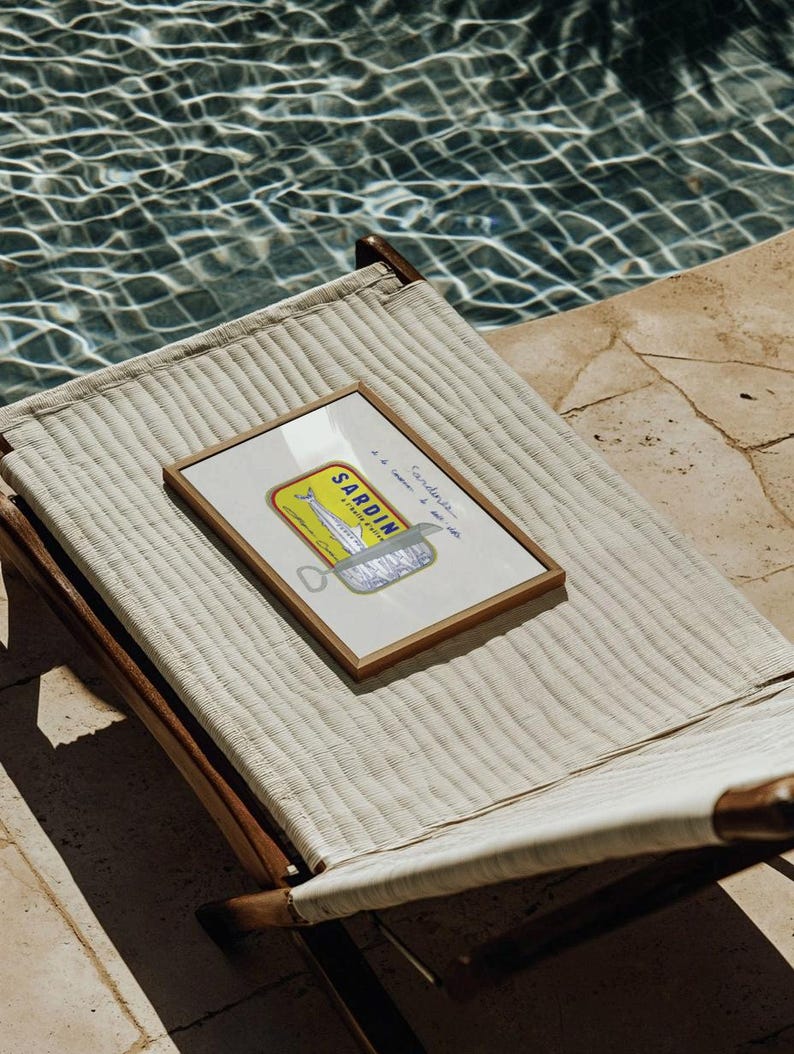 May include: A framed print of a sardine can label rests on a woven lounge chair. The label features a yellow and blue design with the word "SARDINES" and an illustration of a fish. The chair is positioned near a pool.