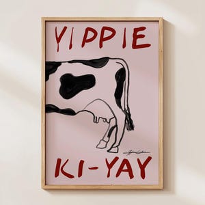 Op de afbeelding: Een ingelijste kunstdruk met een koe-illustratie met zwarte vlekken op een roze achtergrond. De woorden "YIPPIE" en "KI-YAY" staan in het rood boven en onder de koe. Het kunstwerk is rechtsonder gesigneerd.