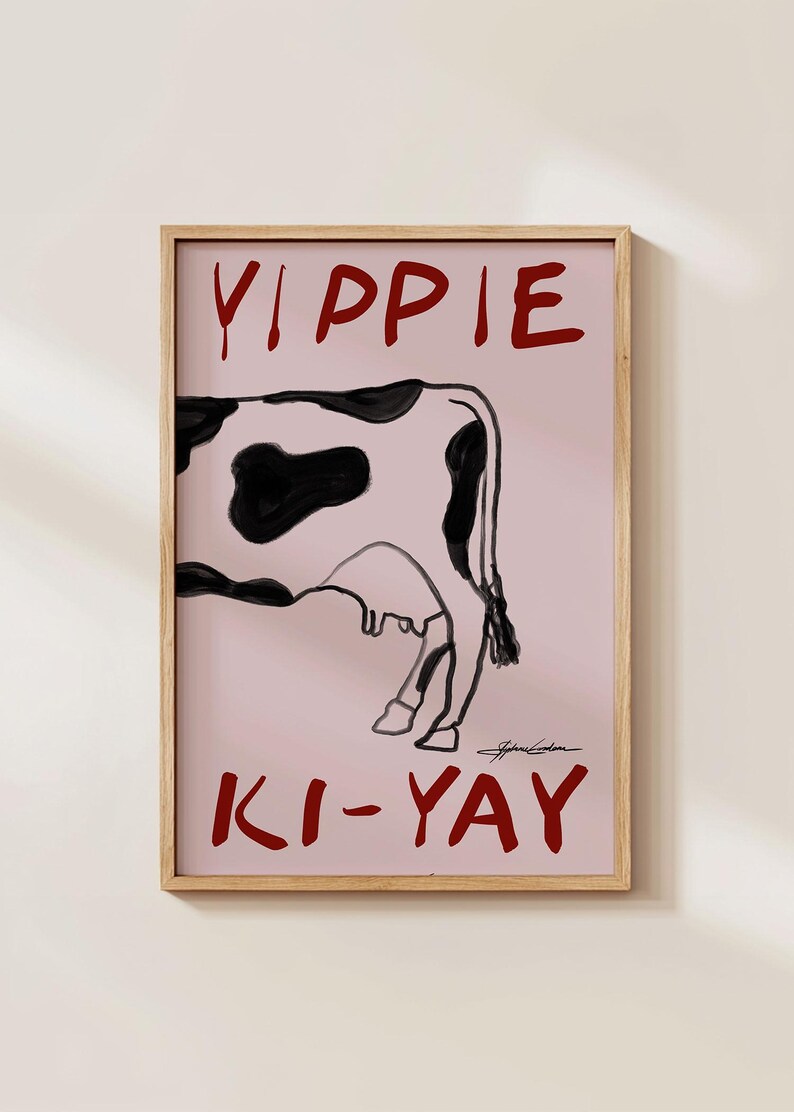Op de afbeelding: Een ingelijste kunstdruk met een koe-illustratie met zwarte vlekken op een roze achtergrond. De woorden "YIPPIE" en "KI-YAY" staan in het rood boven en onder de koe. Het kunstwerk is rechtsonder gesigneerd.