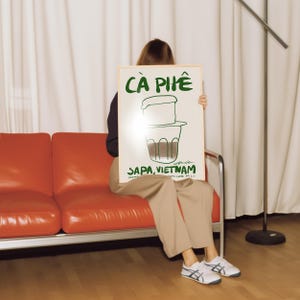 May include: A framed poster with the words "C&Agrave; PH&Ecirc;" in green, along with a drawing of a Vietnamese coffee filter. The text "SAPA, VIETNAM" is also visible. The poster is held by a person sitting on an orange couch.