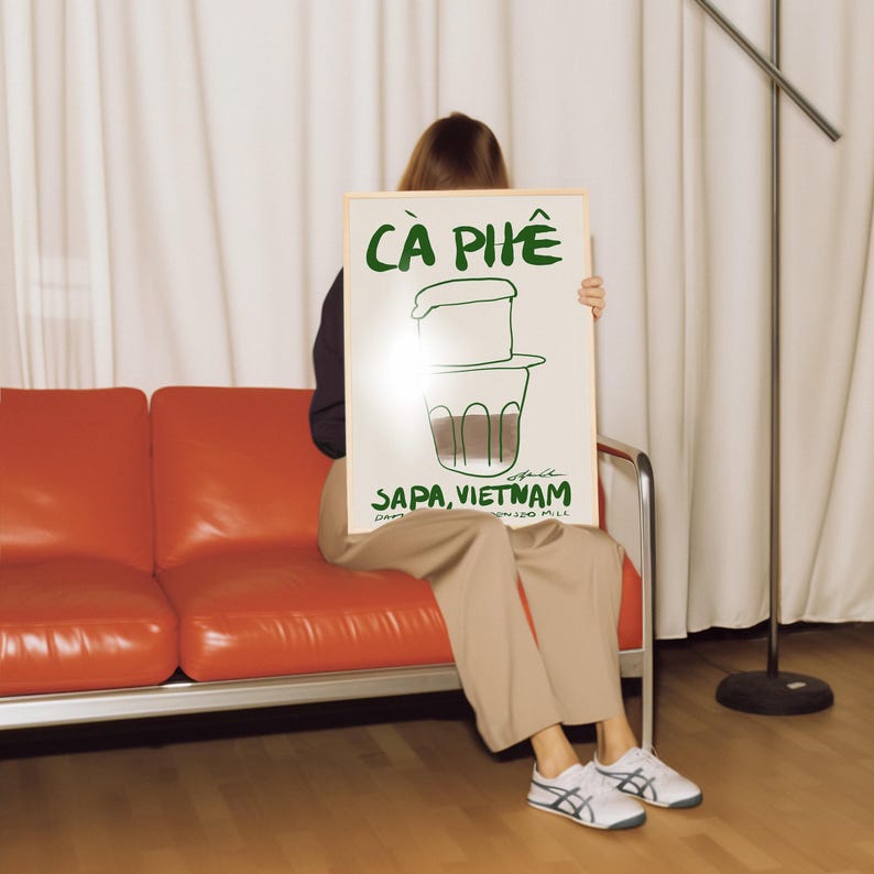 May include: A framed poster with the words "C&Agrave; PH&Ecirc;" in green, along with a drawing of a Vietnamese coffee filter. The text "SAPA, VIETNAM" is also visible. The poster is held by a person sitting on an orange couch.