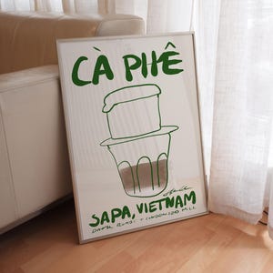 Framed typography poster featuring drink illustration of Vietnamese coffee and bold handwritten lettering by KunstbyStephie, styled in a contemporary minimalist living room with beige leather sofa and linen curtains.