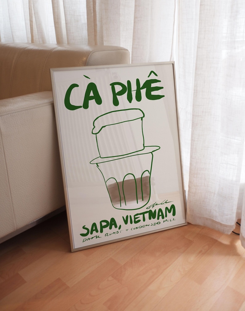 Framed typography poster featuring drink illustration of Vietnamese coffee and bold handwritten lettering by KunstbyStephie, styled in a contemporary minimalist living room with beige leather sofa and linen curtains.