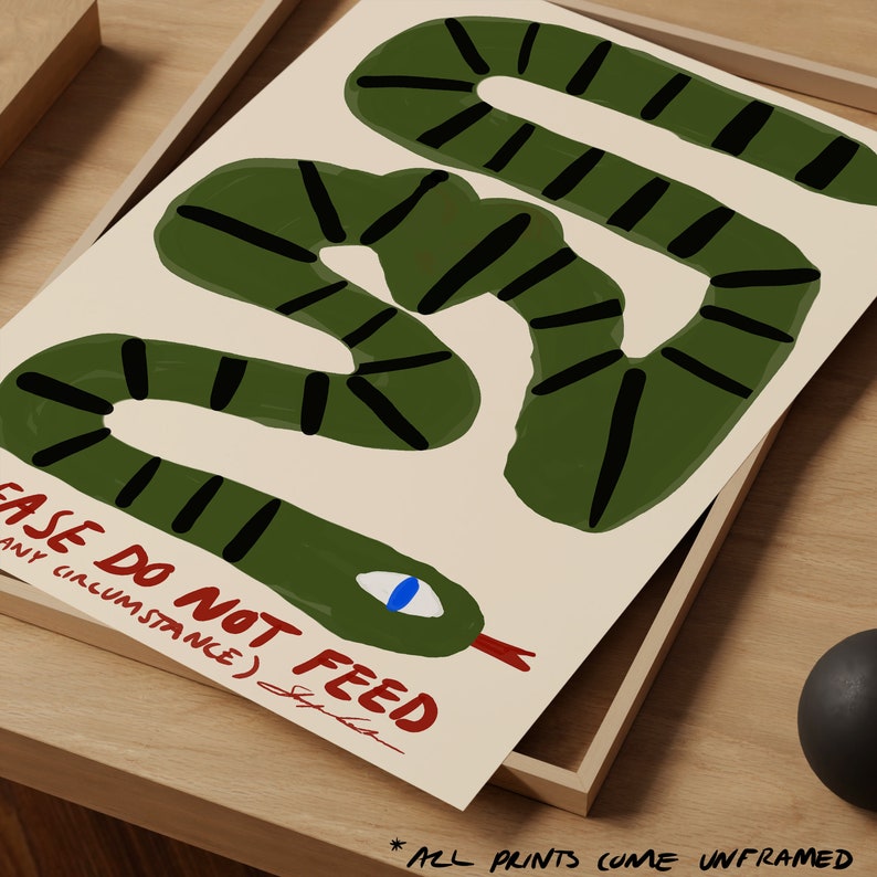 May include: An unframed art print featuring a green snake illustration with black stripes on a cream background. The print includes the text "PLEASE DO NOT FEED" in red. The print is displayed in a wooden frame.