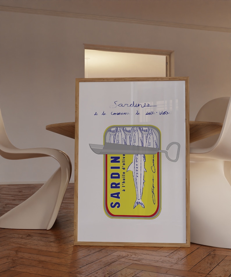 Chic Hand-drawn minimalist kitchen and bar cart print "Sardines" art print poster by KunstbyStephie. A framed print of a hand-drawn illustration of a sardine can with the text 'Sardines - de la Conserverie de la Belle-Iloise' above the can. The can is yellow with a blue label that says 'SARDIN à l'huile d'olive' and a blue drawing of a sardine. The can is open with a silver can opener on top. The print is on a white background and is framed in a light brown wood frame.