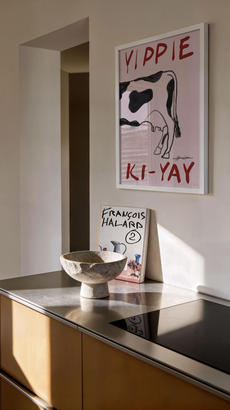 Op de afbeelding: Een ingelijst kunstwerk met een koe-illustratie en het woord "YIPPIE" in het rood. Daaronder staat het woord "KI-YAY" ook in het rood. Een stenen kom staat op een roestvrijstalen aanrechtblad, naast een boek met de titel "FRANCOIS HALARD".