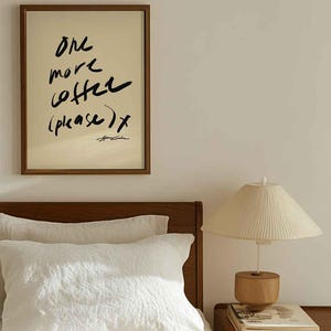 Puede incluir: Un cuadro enmarcado con el texto "One more coffee (please) x" en escritura negra. El cuadro cuelga sobre una cama con ropa de cama blanca, un cabecero de madera y una l&aacute;mpara de noche con una pantalla color crema.