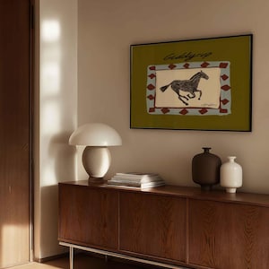 Puede incluir: Una obra de arte enmarcada con una ilustraci&oacute;n de un caballo corriendo y el texto "Giddy Up" sobre un fondo verde. La obra de arte est&aacute; encima de un mueble de madera con una l&aacute;mpara blanca, libros y jarrones decorativos.