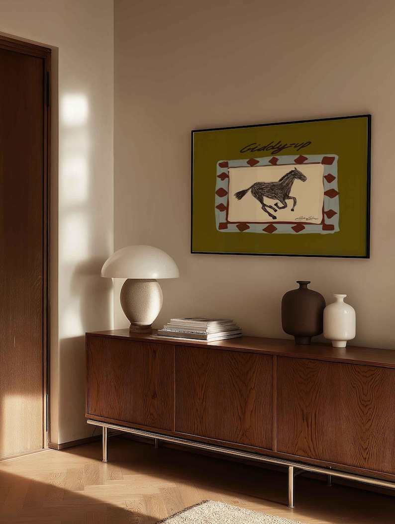 Puede incluir: Una obra de arte enmarcada con una ilustraci&oacute;n de un caballo corriendo y el texto "Giddy Up" sobre un fondo verde. La obra de arte est&aacute; encima de un mueble de madera con una l&aacute;mpara blanca, libros y jarrones decorativos.