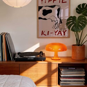 Op de afbeelding: Een retro woonkamer met een houten dressoir, een platenspeler en een oranje paddenstoellamp. Een poster met een koeienafbeelding en de woorden "Yippie Ki-Yay" hangt aan de muur. Een grote papieren lantaarn hangt aan het plafond.