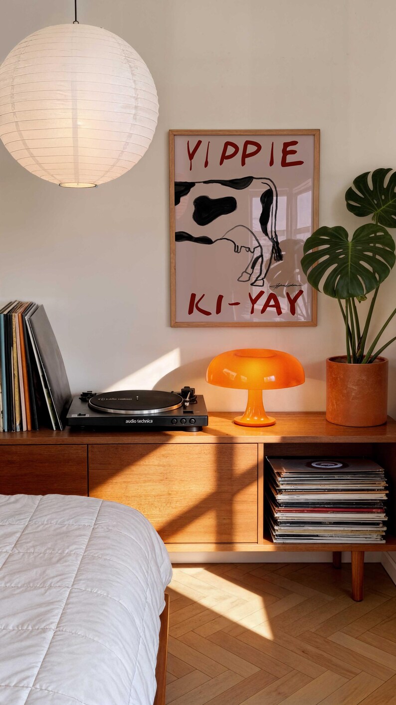 Op de afbeelding: Een retro woonkamer met een houten dressoir, een platenspeler en een oranje paddenstoellamp. Een poster met een koeienafbeelding en de woorden "Yippie Ki-Yay" hangt aan de muur. Een grote papieren lantaarn hangt aan het plafond.