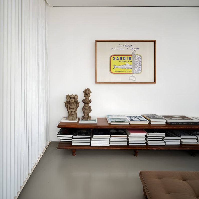 May include: A modern interior featuring a wooden shelf filled with books and decorative items. Above the shelf, a framed artwork depicts a sardine can. The room has white walls, a gray floor, and a brown leather ottoman.