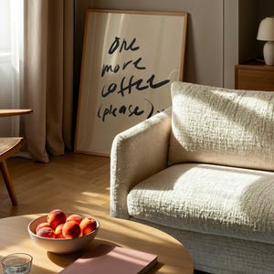 Puede incluir: Escena de sal&oacute;n con una mesa de centro redonda de madera, un sof&aacute; beige y un cuadro enmarcado con el texto "one more coffee (please)." Un cuenco de melocotones y un libro rosa est&aacute;n sobre la mesa.