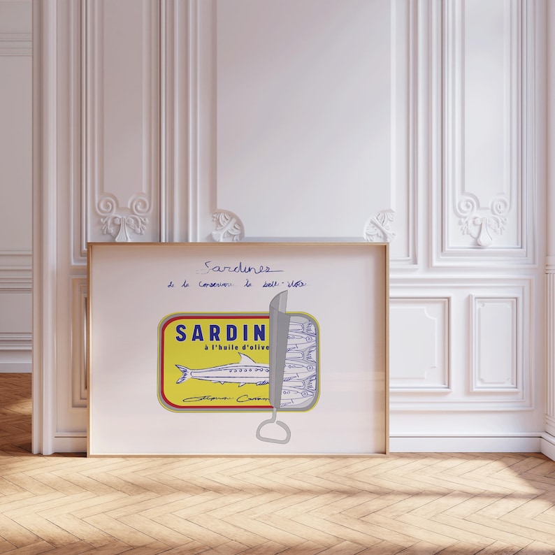 May include: A framed print featuring a sardine can illustration. The can is yellow with blue and red text, and a drawing of a fish. The text "Sardines" is handwritten in blue ink above the can.