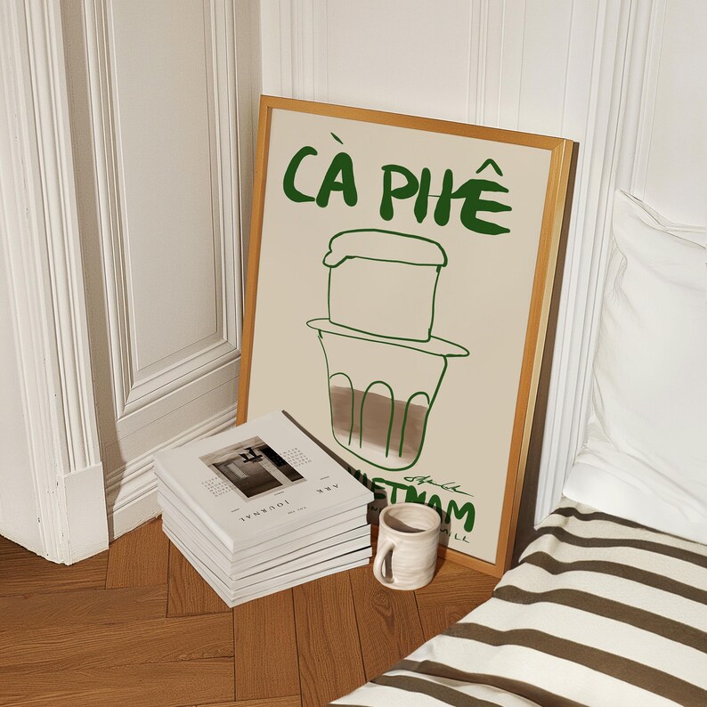 May include: A framed art print with the words "C&Agrave; PH&Ecirc;" in green, depicting a Vietnamese coffee filter. A stack of journals and a small ceramic mug sit on the wooden floor. The print is in a light wood frame.