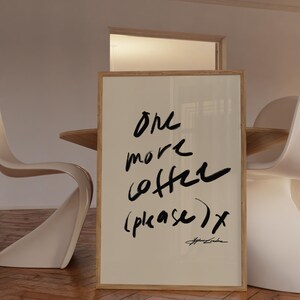 Puede incluir: Una impresi&oacute;n art&iacute;stica enmarcada con las palabras "one more coffee (please) x" en escritura negra. La impresi&oacute;n tiene un fondo beige claro y un marco de madera natural. La obra de arte se exhibe en una habitaci&oacute;n con sillas blancas y una mesa de madera.