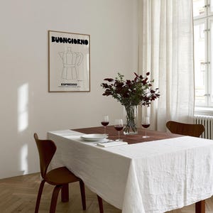 May include: A framed wall art print featuring a black line drawing of a moka pot with the text BUONGIORNO and ESPRESSO in bold black lettering. The print is displayed on a plain wall above a dining table set with a white tablecloth, wine glasses, and a vase.