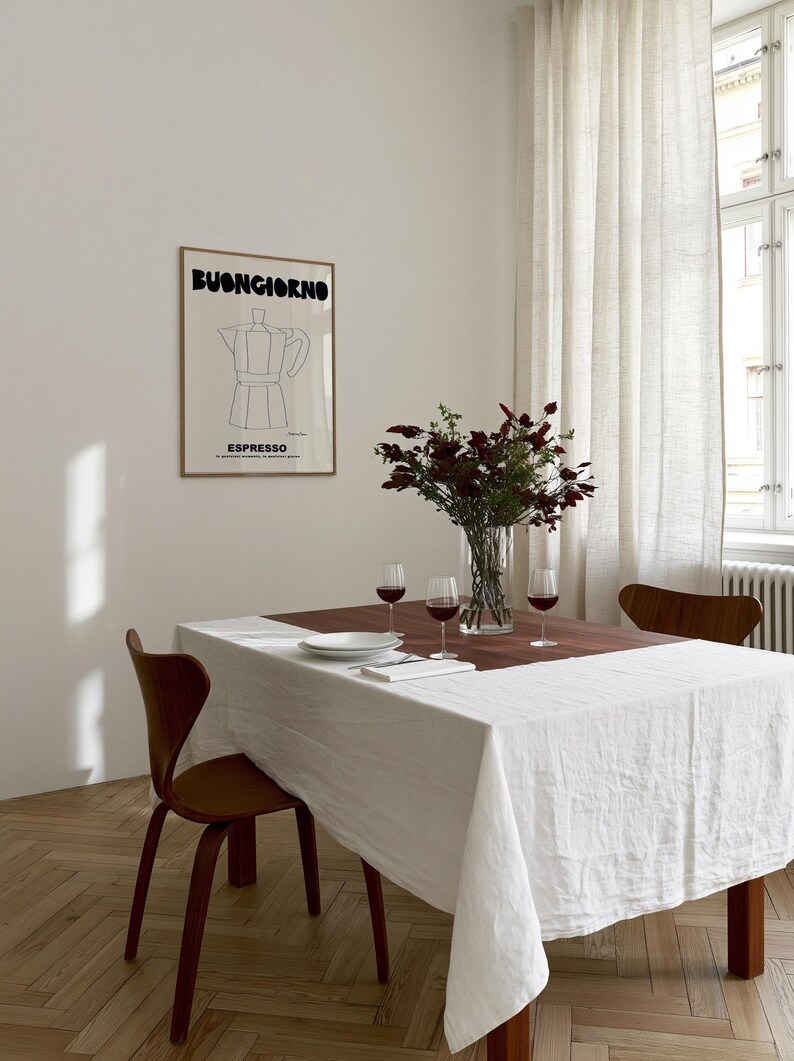 May include: A framed wall art print featuring a black line drawing of a moka pot with the text BUONGIORNO and ESPRESSO in bold black lettering. The print is displayed on a plain wall above a dining table set with a white tablecloth, wine glasses, and a vase.