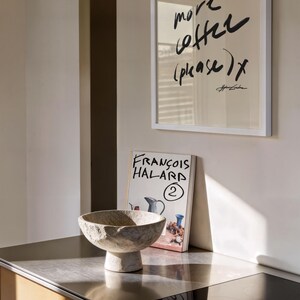 Puede incluir: Un marco blanco con las palabras "one more coffee (please) x" en escritura negra cuelga sobre una encimera. Un cuenco de piedra y un libro titulado "FRANCOIS HALARD" est&aacute;n sobre la encimera. Los armarios de la cocina son de madera.