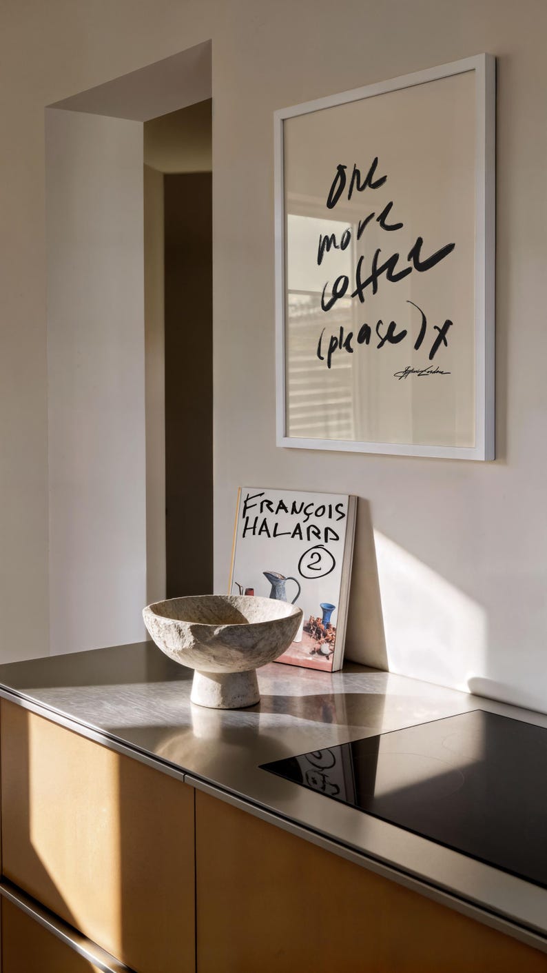Puede incluir: Un marco blanco con las palabras "one more coffee (please) x" en escritura negra cuelga sobre una encimera. Un cuenco de piedra y un libro titulado "FRANCOIS HALARD" est&aacute;n sobre la encimera. Los armarios de la cocina son de madera.