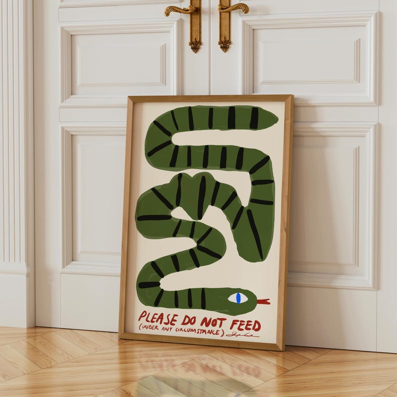 May include: A framed art print depicting a green snake with black stripes on a cream backdrop. The artwork includes the text "PLEASE DO NOT FEED" in red, with additional text below. The snake has a blue eye.
