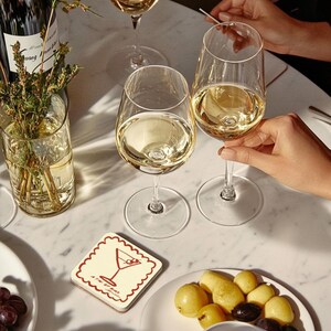May include: A table setting with wine glasses filled with white wine. A coaster with a martini illustration and a plate of olives and potatoes are visible. A bottle of wine and a vase with greenery are also present.