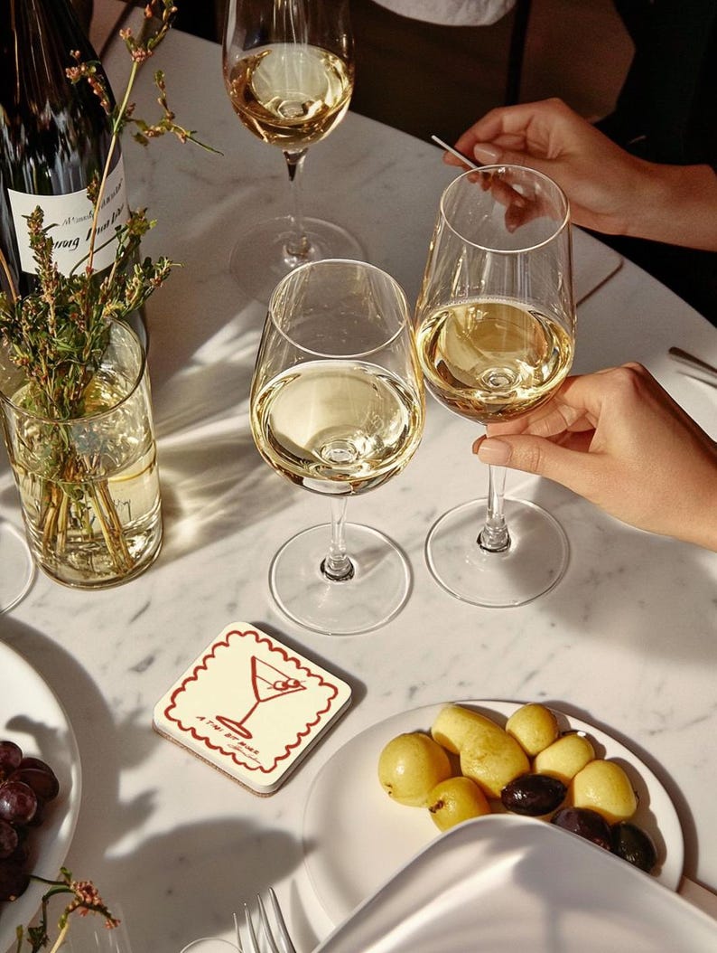 May include: A table setting with wine glasses filled with white wine. A coaster with a martini illustration and a plate of olives and potatoes are visible. A bottle of wine and a vase with greenery are also present.