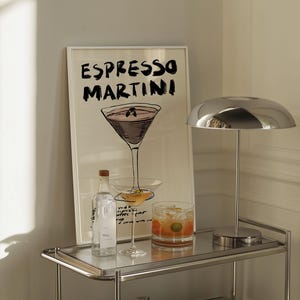 May include: An "Espresso Martini" art print in a white frame, a chrome lamp, a bottle, a cocktail glass, and a rocks glass with a cocktail on a chrome bar cart. The art print features a sketch of a martini glass.