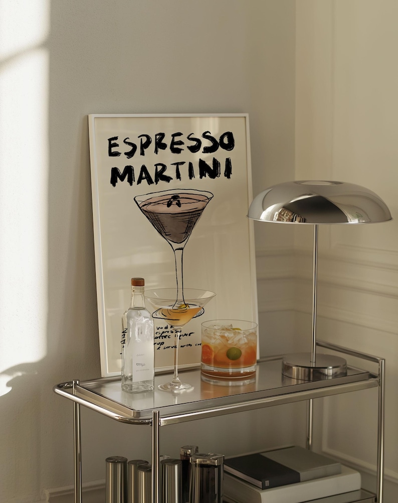 May include: An "Espresso Martini" art print in a white frame, a chrome lamp, a bottle, a cocktail glass, and a rocks glass with a cocktail on a chrome bar cart. The art print features a sketch of a martini glass.