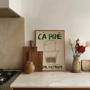 May include: A framed poster with the words "C&Agrave; PH&Ecirc;" in green, depicting a Vietnamese coffee brewing setup. The poster is on a kitchen counter with wooden cutting boards, a vase with flowers, and a small ceramic vase.