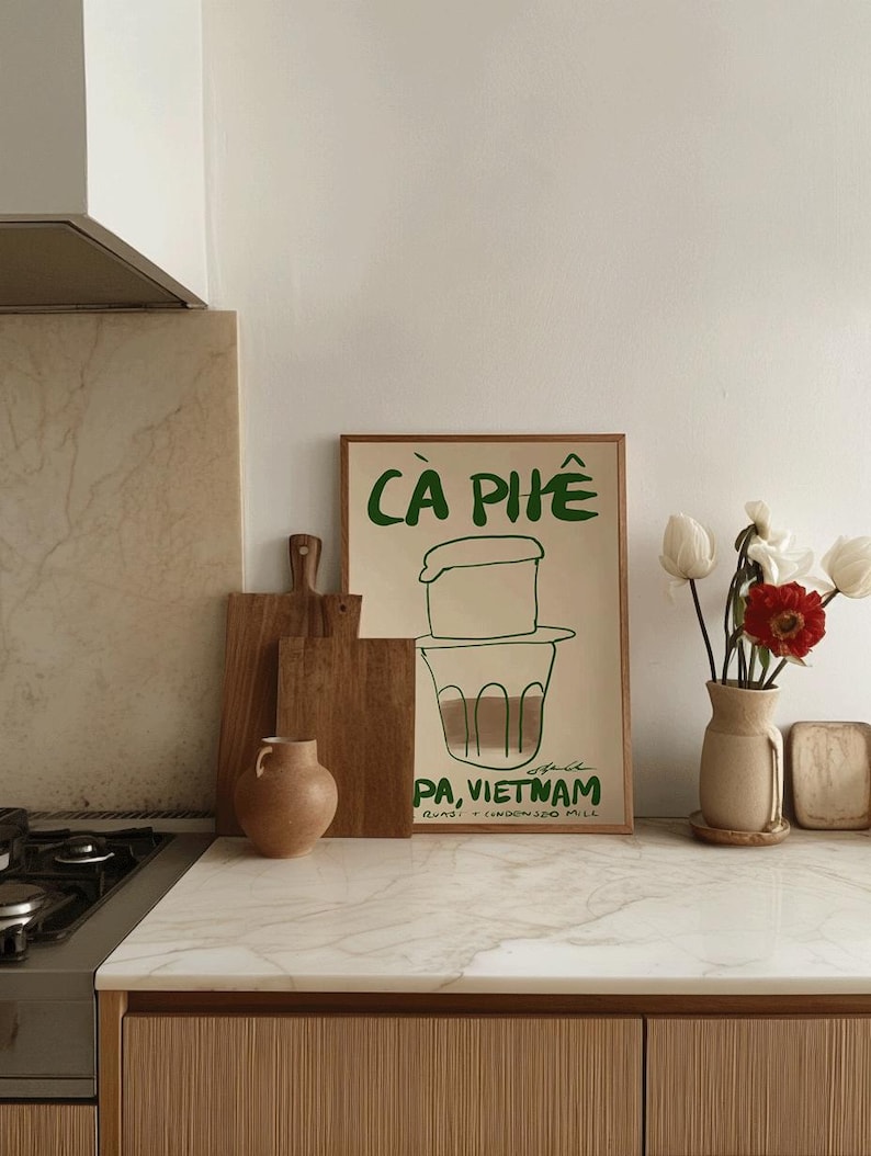 May include: A framed poster with the words "C&Agrave; PH&Ecirc;" in green, depicting a Vietnamese coffee brewing setup. The poster is on a kitchen counter with wooden cutting boards, a vase with flowers, and a small ceramic vase.