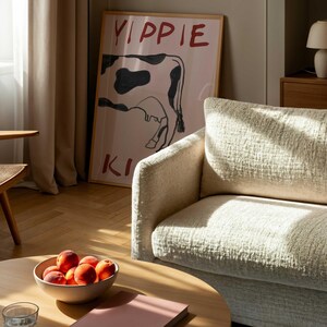 Op de afbeelding: Een woonkamer met een ronde houten salontafel met een kom perziken, een roze boek en een glas water. Een beige bank en een ingelijste poster met een koe-illustratie en de woorden "YIPPIE KI" zijn ook zichtbaar.