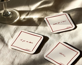 May include: Three square coasters with a white background and a thin, dark red border. Each coaster has a different handwritten phrase in dark red. A wine glass with a clear liquid sits in the background.