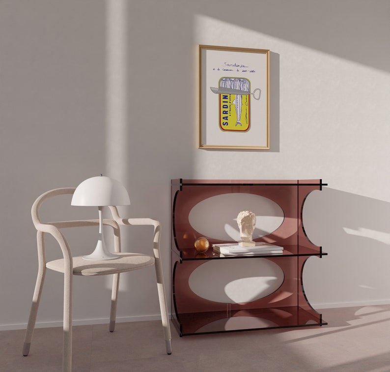 Chic Hand-drawn minimalist kitchen and bar cart print "Sardines" art print poster by KunstbyStephie. A white chair with a curved back and a white lamp with a dome shade. A brown shelf with two shelves and a white bust on the top shelf. A framed print with the words 'Sardines' and a picture of a sardine can with a knife on top of it.
