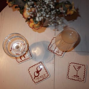 May include: Four square coasters with a scalloped edge and a brown border. One coaster has a martini glass illustration, another has a cocktail shaker, and a third says "Just here for the drinks". A clear glass and a cocktail glass are on the coasters.