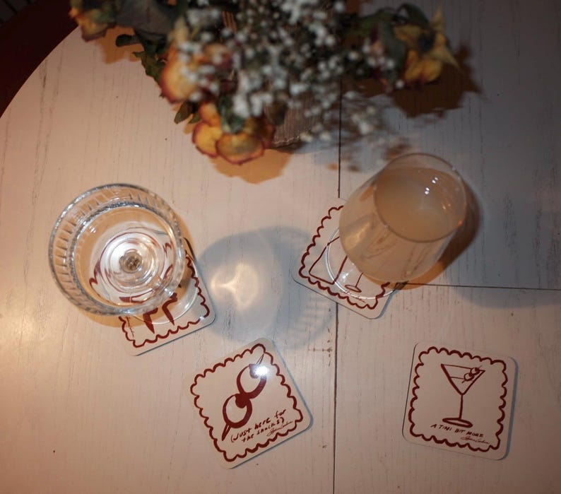 May include: Four square coasters with a scalloped edge and a brown border. One coaster has a martini glass illustration, another has a cocktail shaker, and a third says "Just here for the drinks". A clear glass and a cocktail glass are on the coasters.