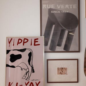 Op de afbeelding: Drie ingelijste kunstwerken hangen aan een witte muur. De grootste toont een koe-illustratie met de rode woorden "Yippie Ki-Yay". Een tweede lijst toont een zwart-wit afbeelding van een stoel. De derde lijst bevat een kleinere, abstracte afbeelding.
