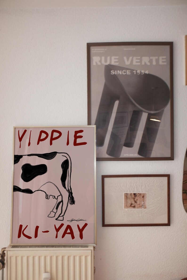 Op de afbeelding: Drie ingelijste kunstwerken hangen aan een witte muur. De grootste toont een koe-illustratie met de rode woorden "Yippie Ki-Yay". Een tweede lijst toont een zwart-wit afbeelding van een stoel. De derde lijst bevat een kleinere, abstracte afbeelding.