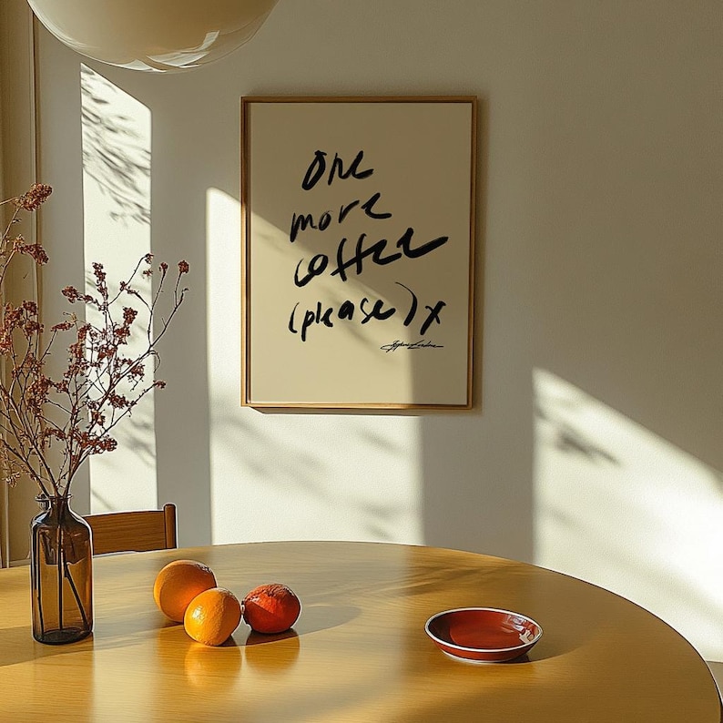 Puede incluir: Un cuadro enmarcado con el texto "One more coffee (please) x" en escritura negra. El cuadro cuelga de una pared color crema. Una mesa de madera tiene naranjas y un plato rojo peque&ntilde;o. Un jarr&oacute;n con ramas secas est&aacute; cerca.