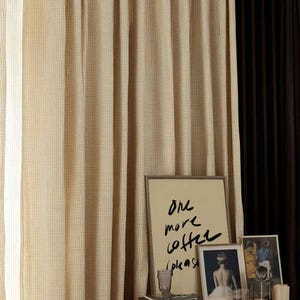 Puede incluir: Cortinas beige y marr&oacute;n a cuadros cuelgan junto a una cortina marr&oacute;n oscuro. Una mesa de madera contiene arte enmarcado, incluida una impresi&oacute;n con el texto "One more coffee please". Otros art&iacute;culos incluyen un vaso, libros y una vela.